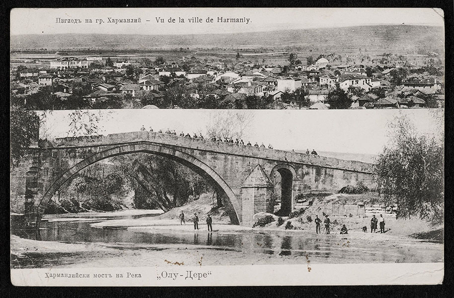 A postcard from 1908. The upper half displays a photograph of Harmanli, showing a small-town setting. The title of the postcard – in both the Cyrillic and Latin scripts – reads: ‘View from the city of Harmanly’. The bottom half shows a photograph of a stone bridge reaching over a shallow river, with a group of twenty townspeople facing the camera.