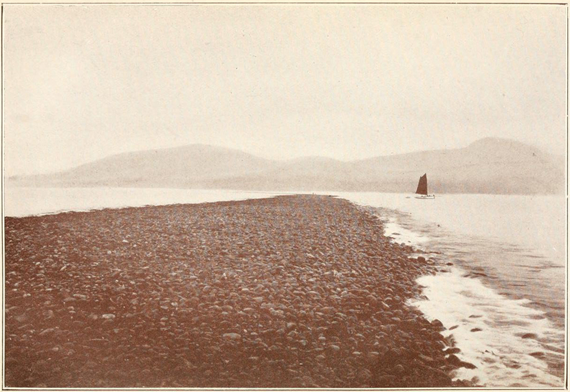 A photograph taken from Cardigan Bay at low tide, looking back to the Welsh coastline, in which the rocky Sarn Badrig appears above the water, and is surrounded by the sea on either side, while a sailing boat approaches the sarn from the right-hand side.