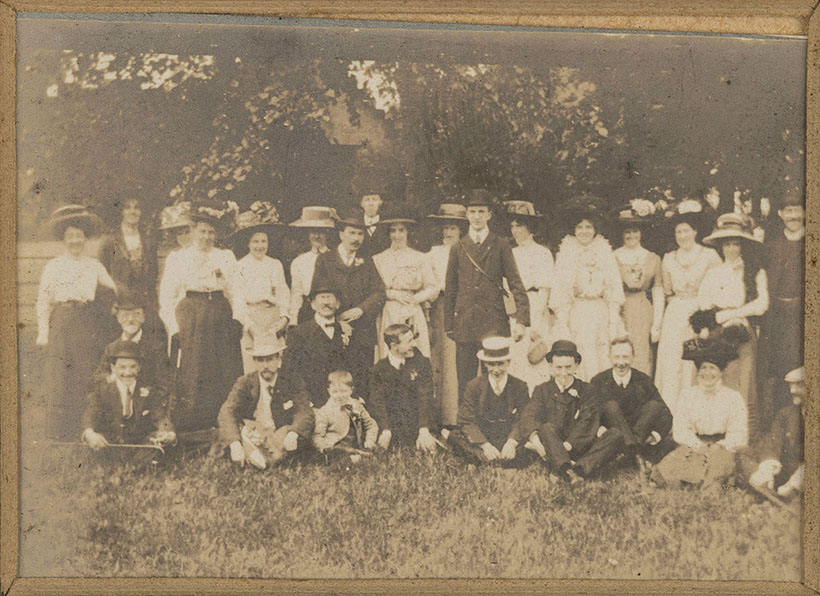 A black-and-white photograph of twenty-eight people – among which thirteen men, fourteen women and a child – in a park. The men wear black suits and hats, and the women wear long skirts, most of them also wearing hats. Eleven people are sitting; the others are standing. All smile spontaneously and face the camera.