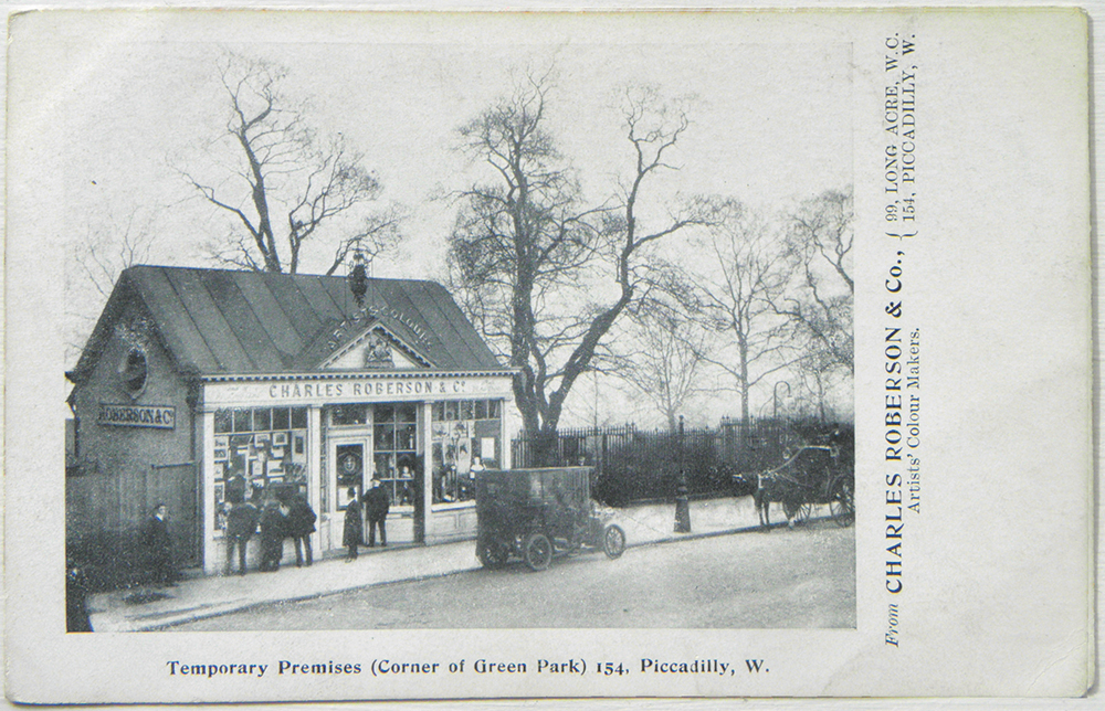 Postcard showing a small, temporary shop on a London street with ‘Charles Roberson & Co.’, ‘Artists’ Colours’ and a royal coat of arms above the door; there are people looking at the goods, mainly pictures, displayed in the windows and a man in a top hat about to open the door; there is a horse-drawn carriage and an early motor vehicle on the street outside and the trees and railings of Green Park can be seen behind and to the right of the shop.