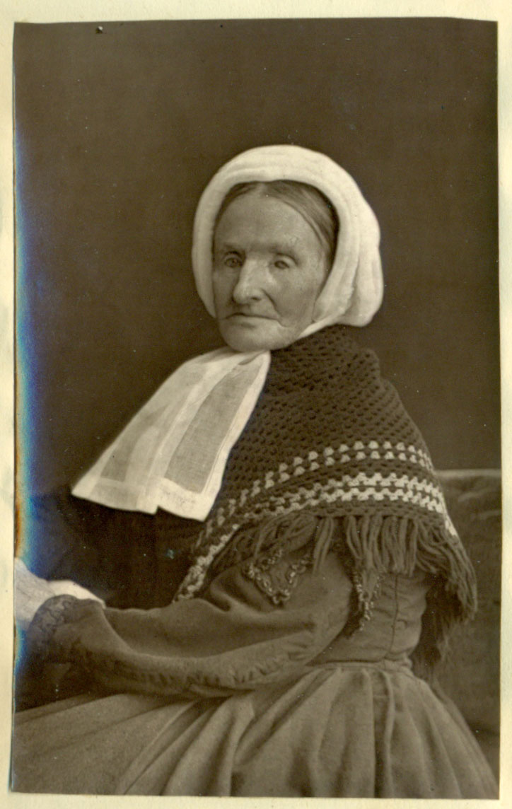 Black and white photograph of an elderly female figure in profile, seated. She wears a heavy, dark-coloured dress, a folded knitted shawl over her shoulders, and a plain white handkerchief around her head with the ends tied loosely at her neck. The accompanying caption reads ‘Mrs Elizabeth Campbell, Aged 72’, including image attribution of ‘J. Valentine, Photo’.