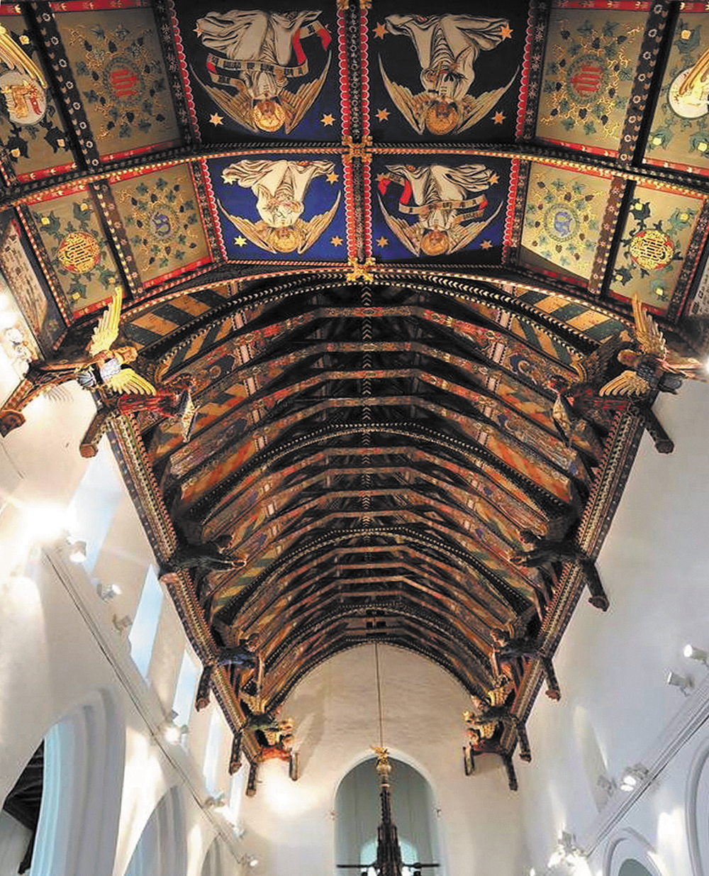 In contrast to the plan, white walls below, the arched ceiling of this church nave is richly and brightly decorated in a medieval style, but executed in the Victorian period, with carved angels along each side where the ceiling meets the walls and with the ceiling itself divided by ribs and beams, with all parts covered in painted and gilded decoration depicting angels, flowers, foliage, animals, geometric patterns and lettered ciphers in green, red, blue, black, white and gold.