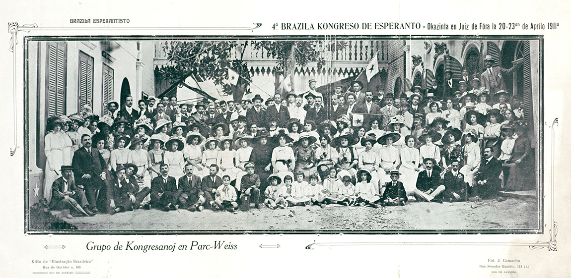 A black-and-white group photograph of where over 100 participants of the Fourth Brazilian Esperanto Congress in in Juiz de Fora, 1911, appear together. The photograph includes men, women and a significant number of children, all wearing formal attires such as suits, long dresses and hats. The people in the picture are lined up in five rows. The background is a school-like building. Three Esperanto flags appear behind the group.