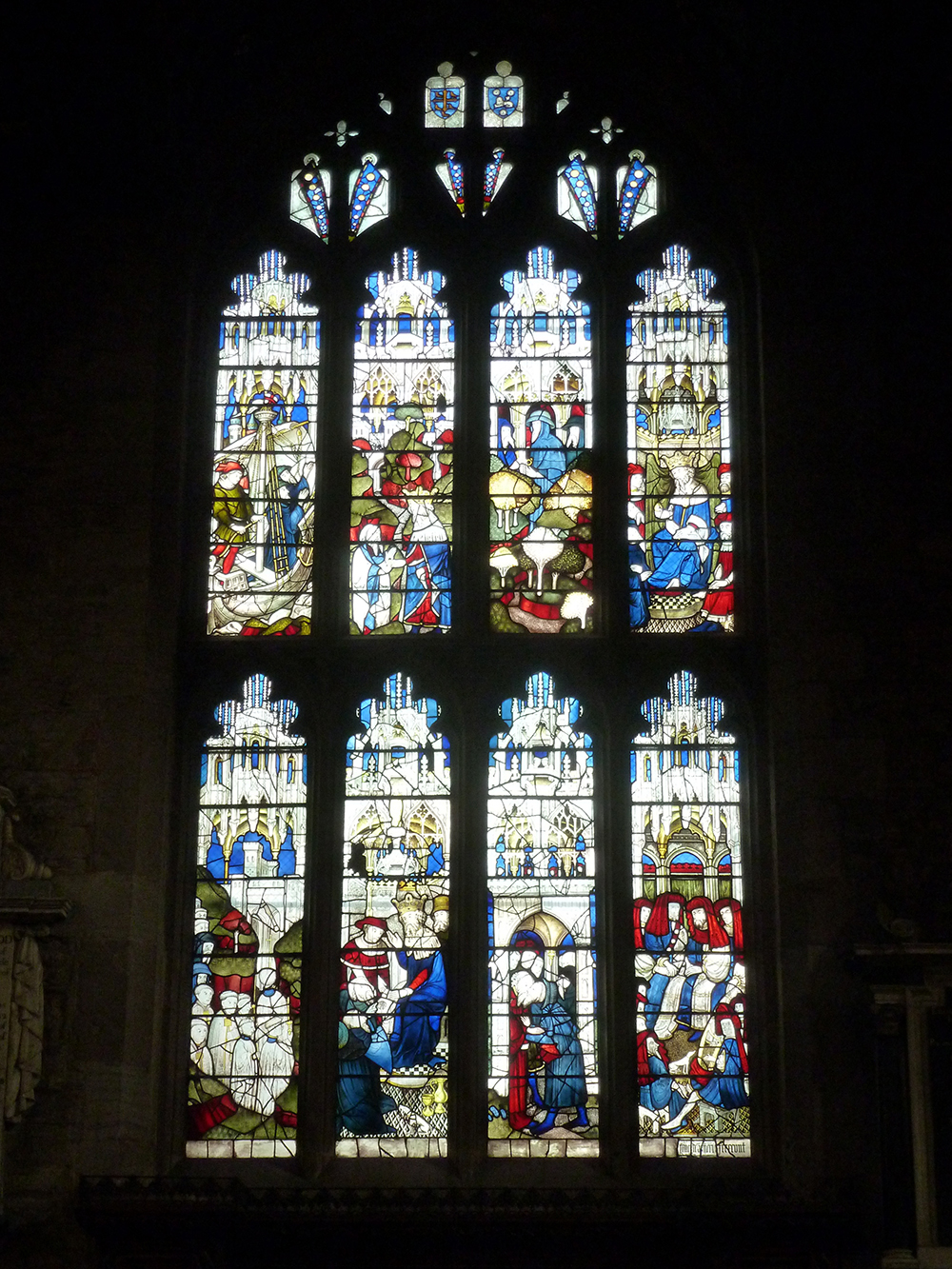 A church window comprising eight stained-glass scenes. The first is of two men on a boat; the second is two men in a field and one man is kneeling. The third comprises three men greeting each other in a field with sheep and trees and the fourth shows a man on a throne surrounded by other men. The fifth scene shows a procession entering a city; the sixth shows two men kneeling in front of a king. In the seventh scene two men kiss each other; the eighth shows a group of men sitting in a circle.