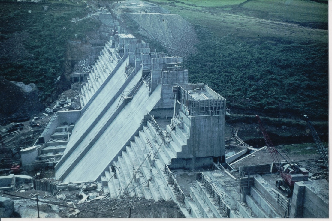 An aerial sidelong view of a partially completed dam wall at Thruscross reservoir. Cranes can be seen in the foreground as some parts of the wall are more complete than ­others.