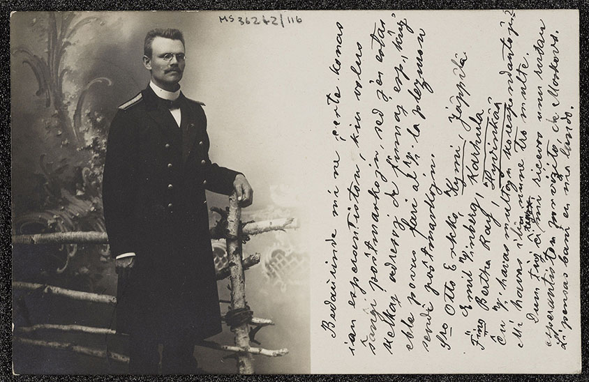 A postcard from 1908 sent by Hugo Salokannel to John Beveridge. The printed image shows Hugo Salokannel, a man wearing a moustache and short hair, standing by a fence and wearing glasses and a black cloak. The handwritten text, in Esperanto, reads: ‘The outfit in which I am dressed represents that of the railway workers in Finland’.