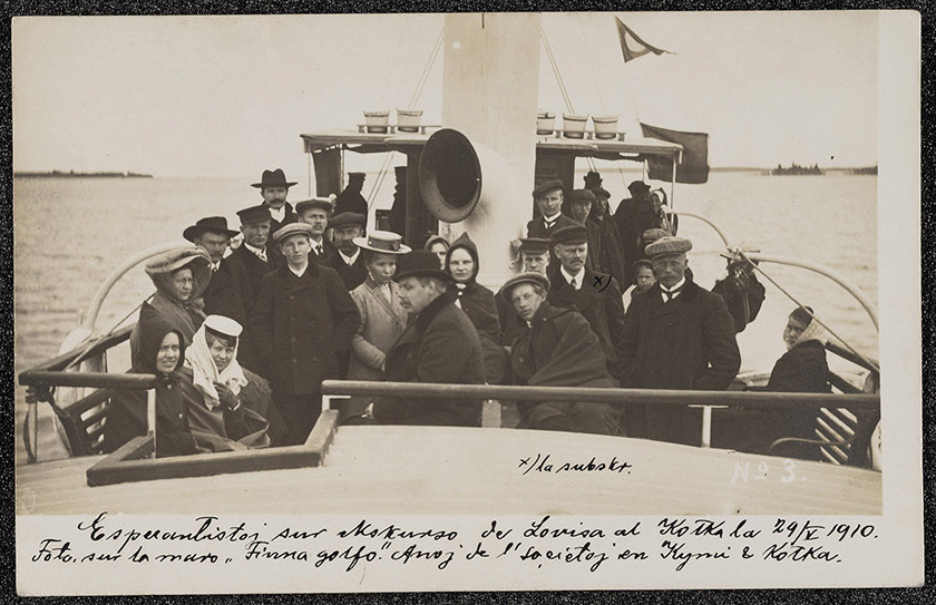 A postcard from 1910 by Hugo Salokannel displaying a photograph of a group of 27 people on a boat, with a short note explaining that these were members of the Esperanto groups of Kymi and Kotka in an outing on the Finnish Gulf in late May 1910. Salokannel marked himself with an ‘X’. The group includes nineteen men, seven women and a child, all wearing black cloaks and most of them covering their heads with a hat or headscarf.