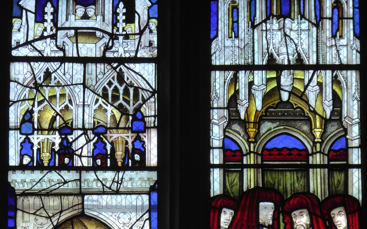 A group of men sit in a circle, holding hands, under an architectural stone canopy. They are all wearing red hoods and gowns that are blue on one side and white and yellow stripped on the other. Next to them, a few men stand underneath another stone canopy, which has two men standing on top of the canopy wearing red hoods.