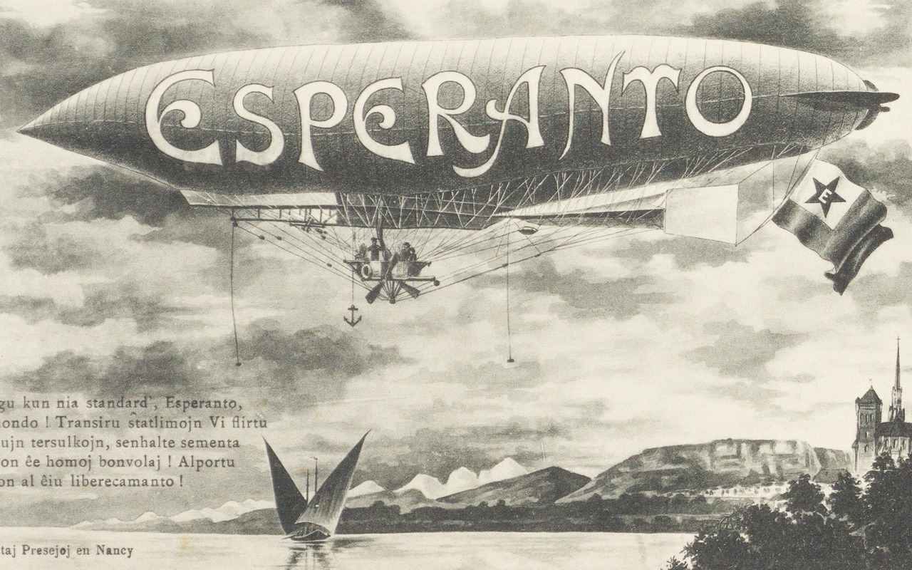 Detail from a postcard, black and white, early 20th century. The postcard, printed in Nancy, France, shows a zeppelin flying over a lake or sea. The zeppelin has the word “Esperanto” printed on the side from front to back. At its tail it carries the green and white Esperanto flag. The background scenery shows a mountainous coastline and a cathedral
